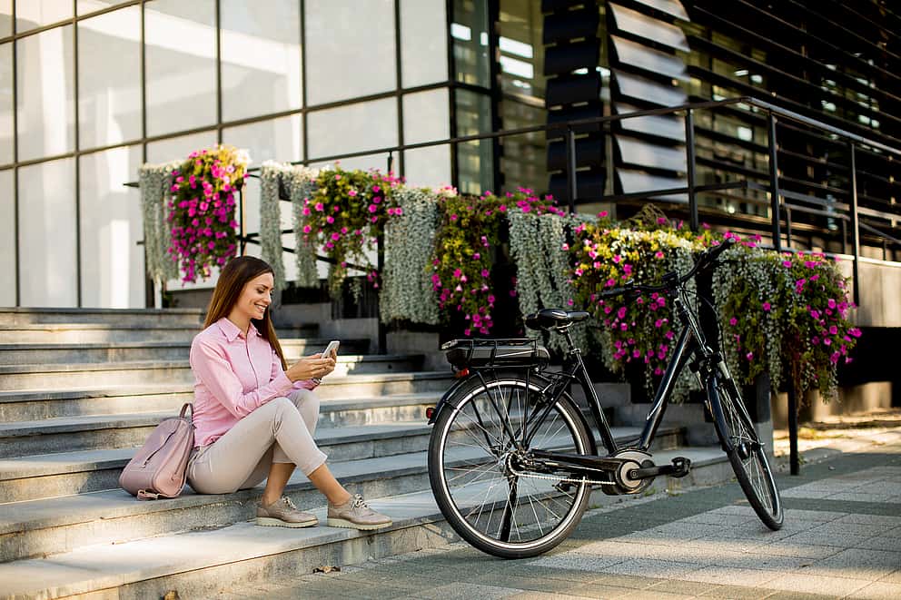 Eine Frau sitzt vor einem Gebäude auf einer Treppe und schaut auf ihr Smartphone, vor ihr steht ein Fahrrad.