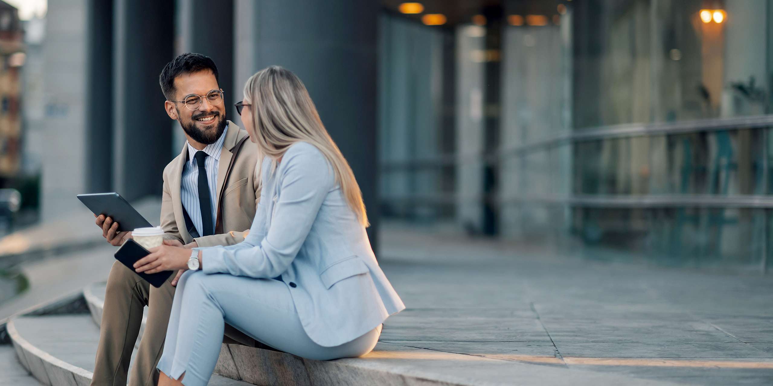 Eine Frau und ein Mann in Business-Kleidung sitzen mit Smartphone und Tablet auf einer Treppe.