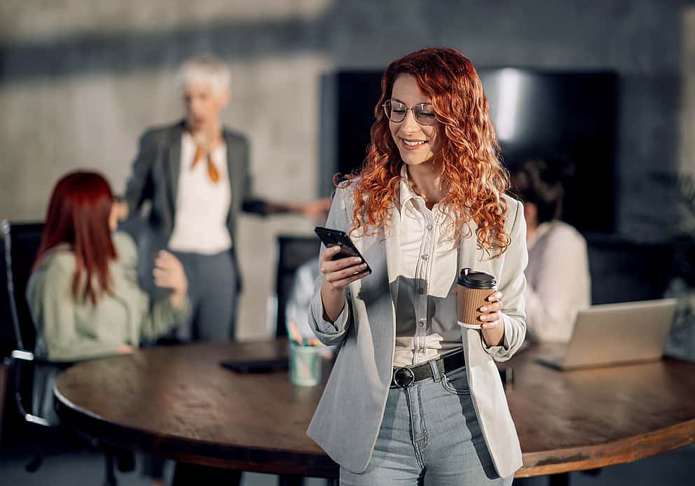 Eine Frau sitzt angelehnt an einem Konferenztisch mit einem Smartphone in der Hand.